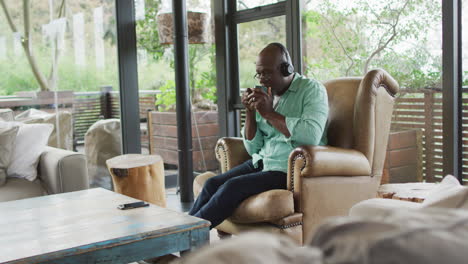 Happy-african-american-senior-man-relaxing-in-armchair,-wearing-headphones-and-drinking-coffee