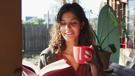 Happy-mixed-race-woman-sitting-drinking-tea-and-reading-book-in-sunny-cottage-kitchen