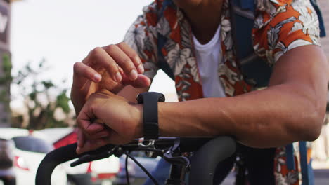 Hombre-Afroamericano-En-La-Ciudad,-Sentado-En-Bicicleta-En-La-Calle-Usando-Smartwatch
