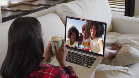 Eine-Afroamerikanische-Frau-Hält-Eine-Kaffeetasse-In-Der-Hand-Und-Führt-Einen-Videoanruf-Auf-Einem-Laptop,-Der-Zu-Hause-Auf-Der-Couch-Sitzt