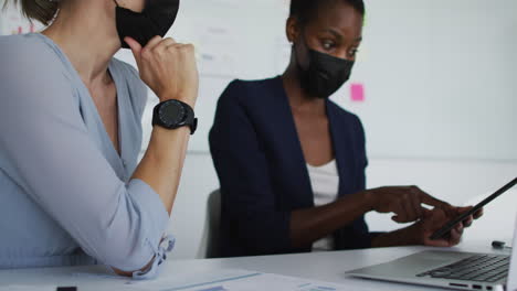 Two-diverse-female-colleagues-wearing-face-mask,-sitting-at-desk-and-talking