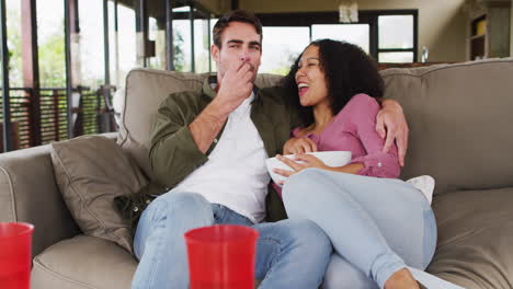 Smiling-mixed-race-couple-eating-popcorn-sitting-together-on-the-couch-at-vacation-home