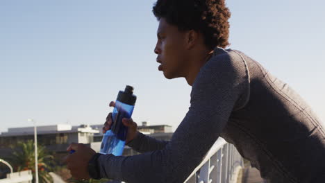 Fit-african-american-man-exercising-in-city,-resting-on-footbridge,-drinking-from-water-bottle