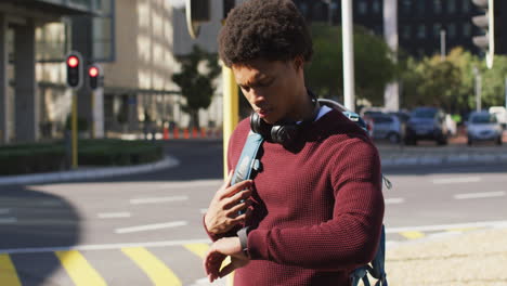 Hombre-Afroamericano-En-La-Ciudad-Revisando-Smartwatch,-Usando-Audífonos-Y-Mochila-Esperando-En-La-Calle