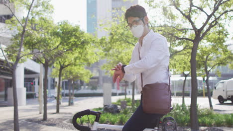 Asiatischer-Mann-Mit-Gesichtsmaske-Und-Smartwatch,-Während-Er-Auf-Seinem-Fahrrad-Auf-Der-Straße-Sitzt