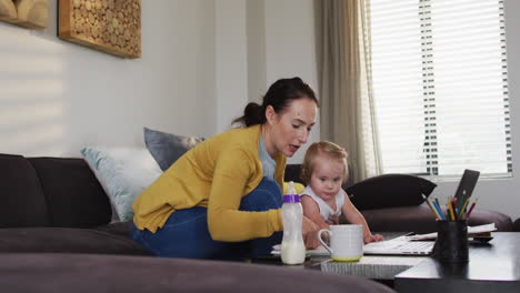Caucasian-mother-holding-her-baby-using-laptop-and-taking-notes-while-working-from-home