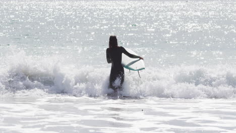 Rear-view-of-mixed-race-woman-walking-into-the-sea-carrying-surfboard