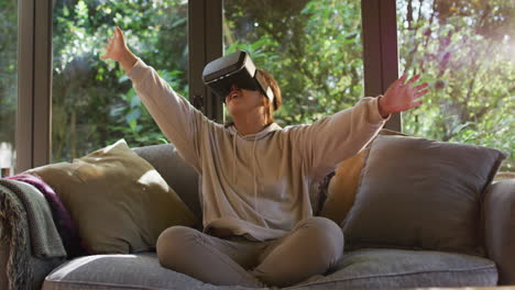 Asian-girl-smiling-and-gesturing-while-wearing-vr-headset-sitting-on-the-couch-at-home