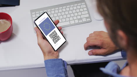 Man-at-desk-holding-smartphone-with-covid-vaccination-certificate,-eu-flag-and-qr-code-on-screen