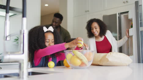 Feliz-Niña-Afroamericana-Desempacando-Comestibles-En-La-Cocina-Con-Sus-Padres-Y-Su-Hermano