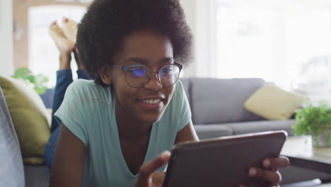 Happy-african-american-teenage-girl-lying-on-sofa,-using-tablet