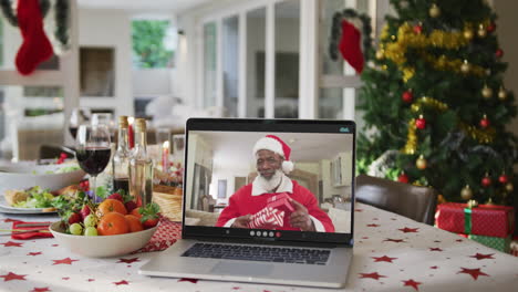 Fröhlicher-Afroamerikanischer-Weihnachtsmann-Auf-Dem-Laptop,-Der-Auf-Dem-Weihnachtstisch-Liegt