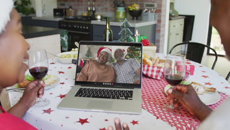 African-american-couple-with-wine-using-laptop-for-christmas-video-call-with-couple-on-screen