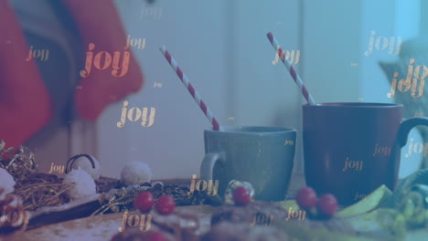 Animation-of-joy-text-falling-over-christmas-decorations-and-mugs-of-hot-chocolate