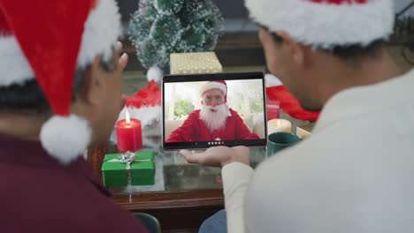 Biracial-father-and-son-with-santa-hats-using-tablet-for-christmas-video-call-with-santa-on-screen