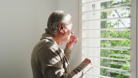 Retired-senior-man-in-cardigan-sweater-looking-out-through-window-blinds-at-home