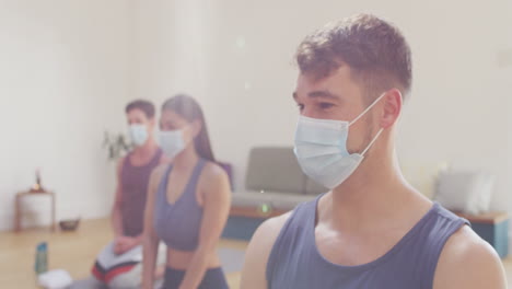Diverse-group-of-men-and-woman-in-face-masks-kneeling-on-mats-at-yoga-class-listening-to-instructor