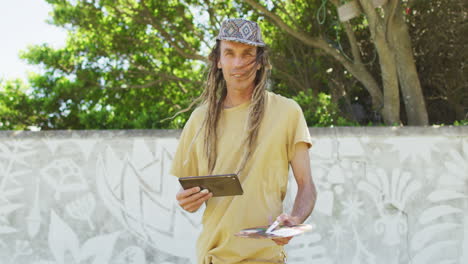 Video-of-smiling-caucasian-man-with-dreadlocks-holding-tablet-and-painter's-palette-by-mural-on-wall