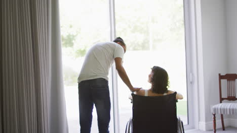 Biracial-woman-in-wheelchair-and-smiling-male-partner-talking-and-looking-out-of-living-room-window