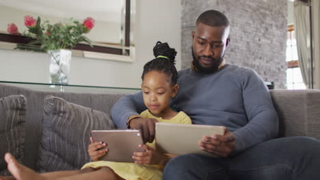Glückliche-Afroamerikanische-Tochter-Und-Vater,-Die-Tablets-Auf-Dem-Sofa-Benutzen