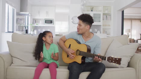 Happy-biracial-father-and-daughter-sitting-on-sofa-playing-guitar