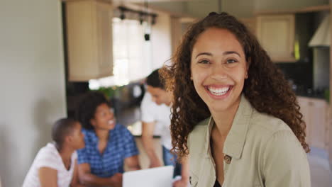 Happy-biracial-woman-standing-in-kitchen-and-looking-at-camera-with-diverse-friends-in-background
