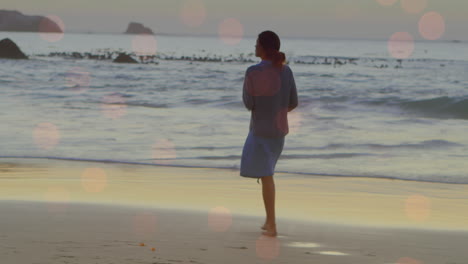 Animation-of-light-spots-over-biracial-woman-walking-on-beach
