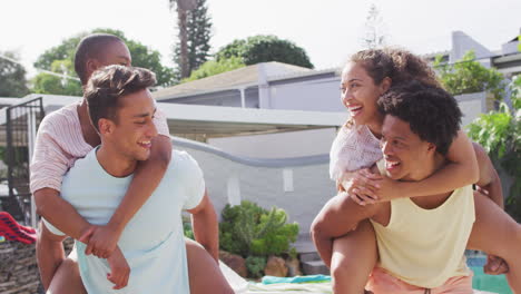 Two-happy-diverse-male-friends-carrying-piggyback-female-friends-in-backyard-at-pool-party