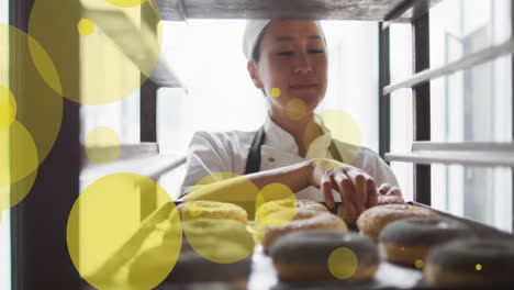 Animation-of-green-spots-over-asian-female-baker-preparing-donuts