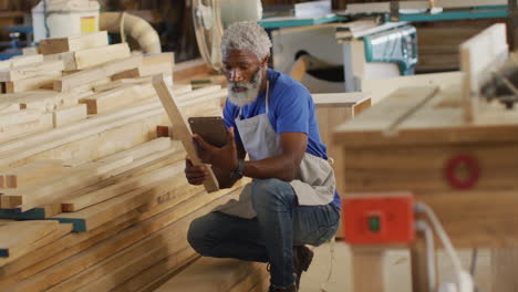 Afroamerikanischer-Zimmermann-Mit-Digitalem-Tablet,-Der-In-Einer-Tischlerei-Ein-Holzbrett-Auswählt