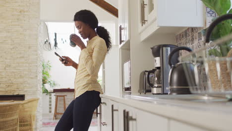 Video-of-african-american-woman-drinking-coffee-and-using-smartphone-in-kitchen