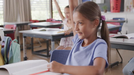 Happy-diverse-schoolgirls-at-desks-raising-hands-in-classroom-at-elementary-school