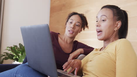 Happy-biracial-lesbian-couple-relaxing-on-couch-together,-using-laptop-and-talking,-in-slow-motion