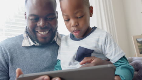 Glücklicher-Afroamerikanischer-Vater,-Der-Mit-Seinem-Sohn-Auf-Dem-Schoß-Auf-Dem-Sofa-Sitzt-Und-Ein-Tablet-Benutzt,-In-Zeitlupe