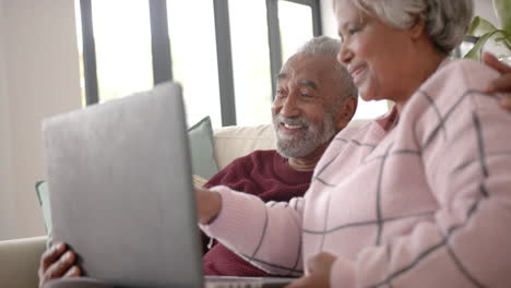 Happy-senior-biracial-couple-sitting-on-sofa-and-using-laptop,-unaltered,-in-slow-motion
