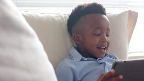 Feliz-Niño-Afroamericano-Tumbado-En-El-Sofá-Usando-Una-Tableta-En-Casa,-Cámara-Lenta