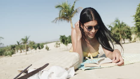 Fröhliche-Hispanische-Frau-Mit-Sonnenbrille,-Die-Am-Strand-Liegt-Und-Ein-Buch-In-Der-Sonne-Liest,-Kopierraum,-Zeitlupe