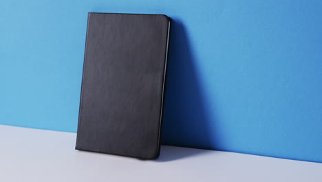 Nahaufnahme-Eines-Geschlossenen-Schwarzen-Buches,-Das-An-Der-Wand-Lehnt,-Mit-Kopierraum-Auf-Blauem-Hintergrund-In-Zeitlupe