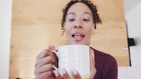 Happy-biracial-woman-making-video-call-talking-and-drinking-coffee-in-living-room,-in-slow-motion