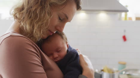 Video-of-caucasian-mother-with-newborn-baby-in-baby-carrier-hugging-in-kitchen