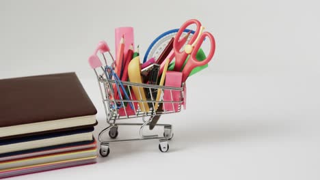 Close-up-of-shopping-trolley-with-school-items-and-stack-of-books-on-grey-background