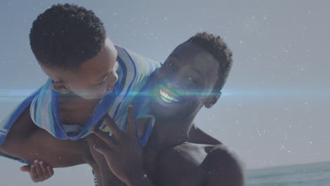Animation-Von-Lichtspuren-über-Einem-Afroamerikanischen-Mann-Und-Seinem-Sohn,-Die-Zusammen-Am-Strand-Spielen