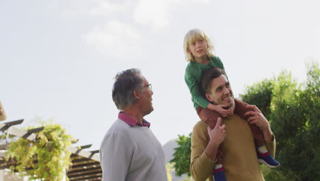 Video-of-happy-caucasian-father-carrying-son-on-shoulders-and-walking-with-grandfather-in-garden