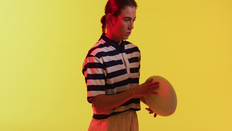 Portrait-of-caucasian-female-rugby-player-with-rugby-ball-over-neon-yellow-lighting