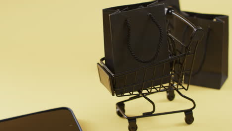 Video-Eines-Smartphones-Mit-Leerem-Bildschirm,-Einkaufstaschen-Und-Einkaufswagen,-Kopierraum-Auf-Gelbem-Hintergrund