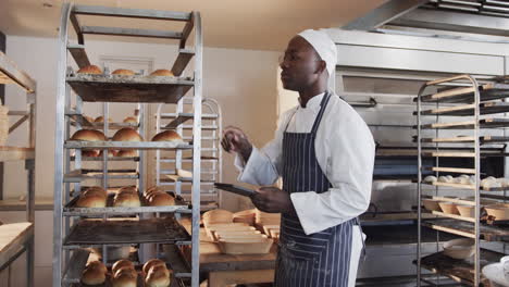 African-american-male-baker-working-in-bakery-kitchen,-counting-rolls-with-tablet-in-slow-motion