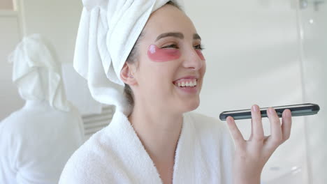 Caucasian-woman-in-bathrobe-with-towel-on-head-and-eye-masks-using-phone-in-bathroom,-slow-motion