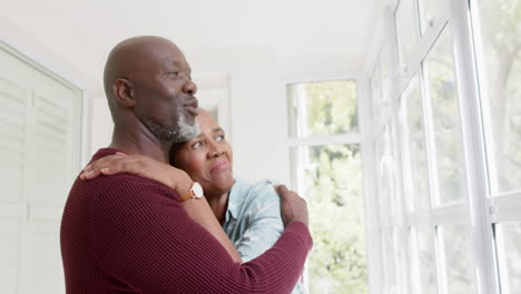 Glückliches-älteres-Afroamerikanisches-Paar,-Das-Sich-Zu-Hause-Umarmt-Und-Aus-Dem-Fenster-Schaut,-Zeitlupe