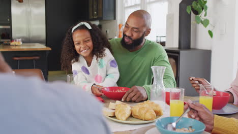 Video-of-happy-african-american-daughter-laughing-and-feeding-father-at-family-breakfast-table