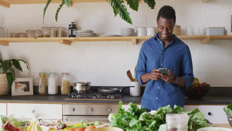 Feliz-Hombre-Afroamericano-Cocinando-La-Cena-En-La-Cocina,-Usando-Un-Teléfono-Inteligente